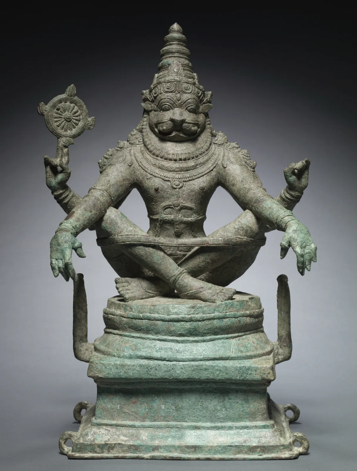 A green-patinated bronze figure seated cross-legged on a stepped base, with a lion's face, a tall crown and four arms. A band loops around his raised knees. Two thin posts rise from the base on either side of him.