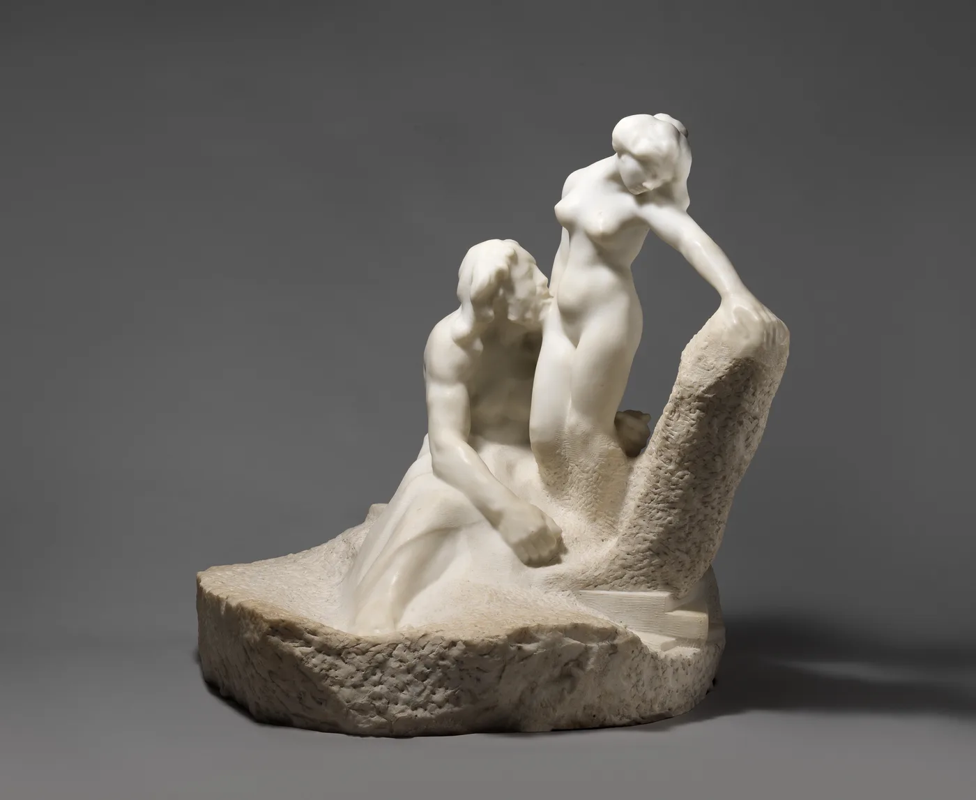 A white marble sculpture on a rough-cut stone base. A bearded, seated man leans his head against a standing nude woman, who rests one hand on a tall block of uncut stone. Her lower legs merge into the rough stone.